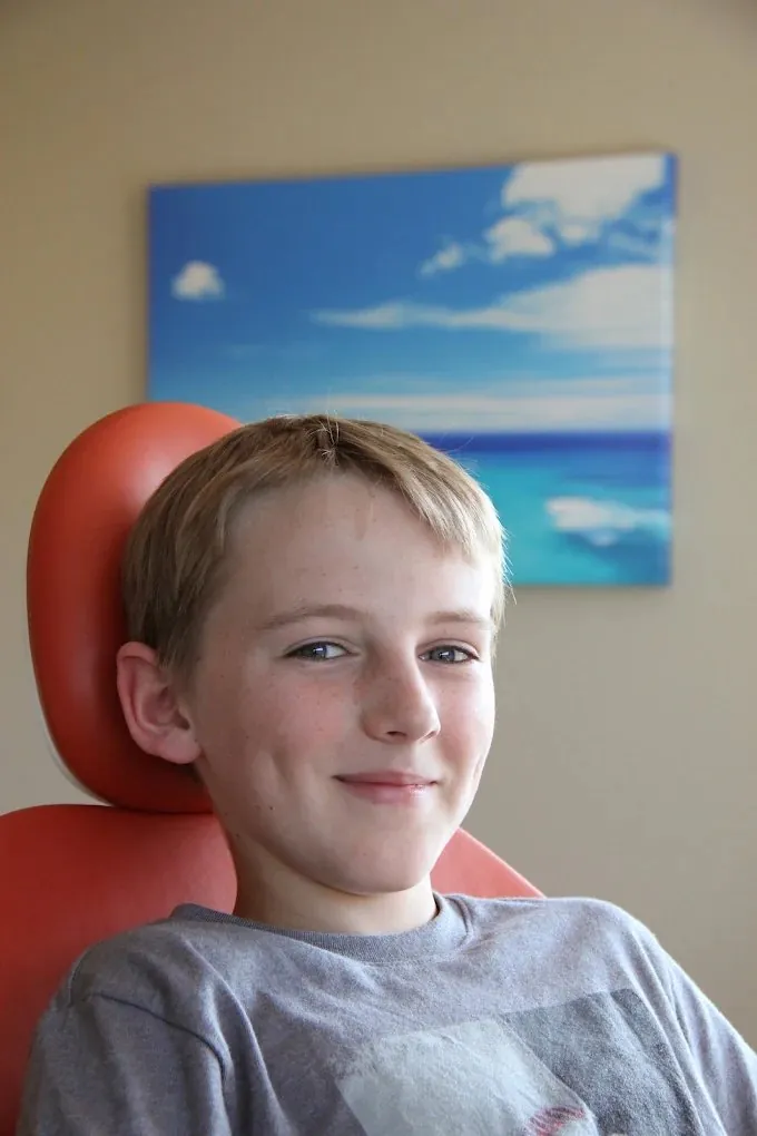 A smiling boy sitting in an orange dental chair under a beach painting